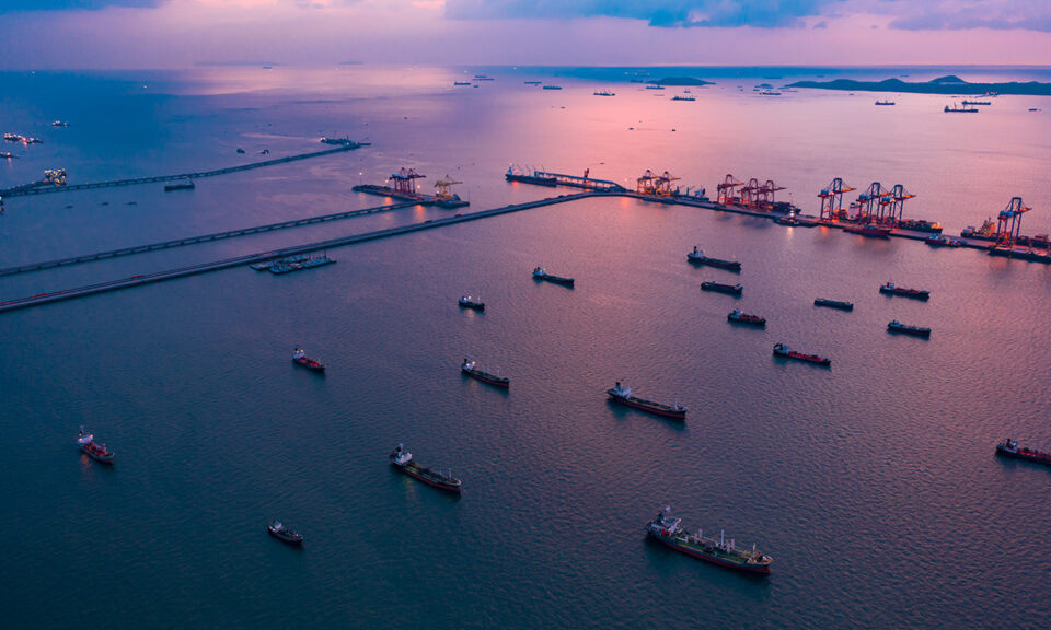 Flera fraktfartyg ligger förtöjda i vattnet och väntar. Solen har gått ner och himlen lyser i rosa.