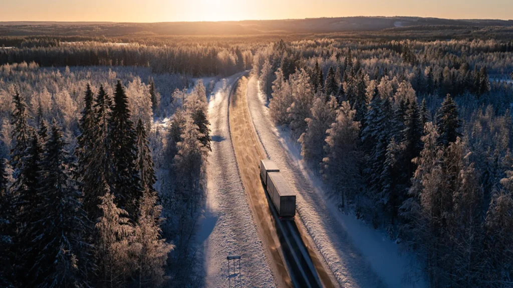 En lastbil kör på en landsväg genom skogen. Det är vinter och solen håller på att gå ner.