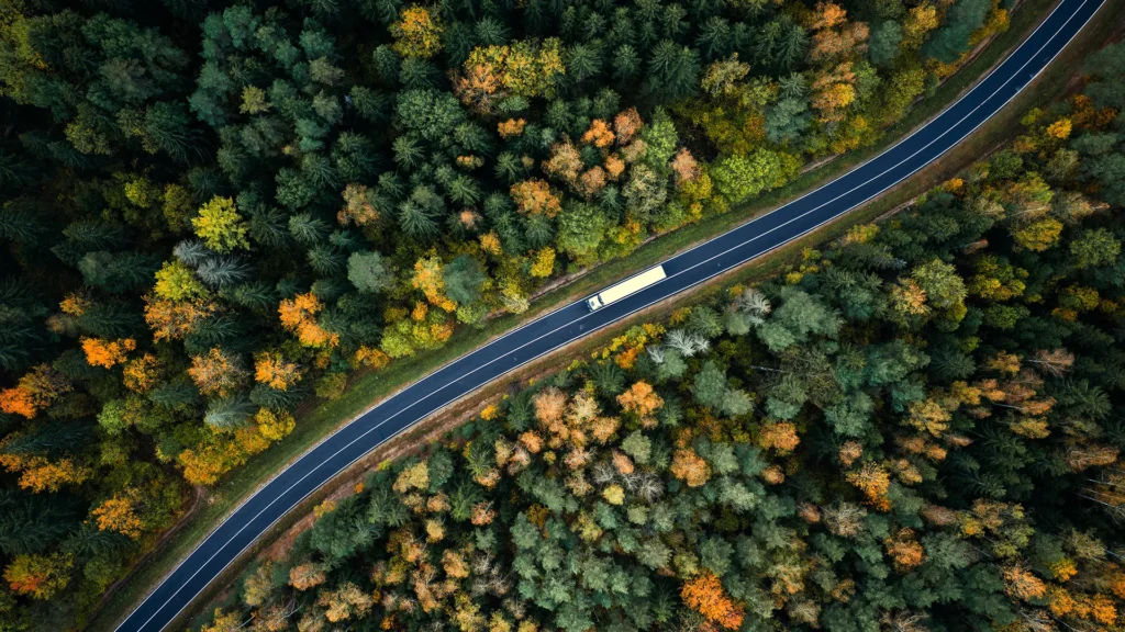 Bilden är tagen ovanifrån. Den visar en lastbil som kör på en väg omgärdad av skog. Det är höst.