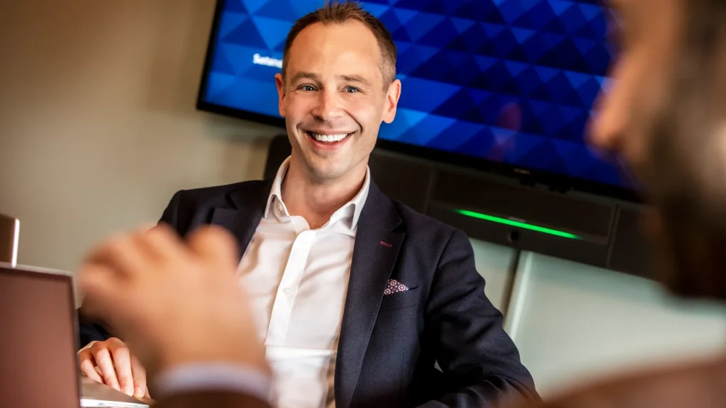 En man sitter i ett konferensrum på ett kontor. Han ler mot en annan man.