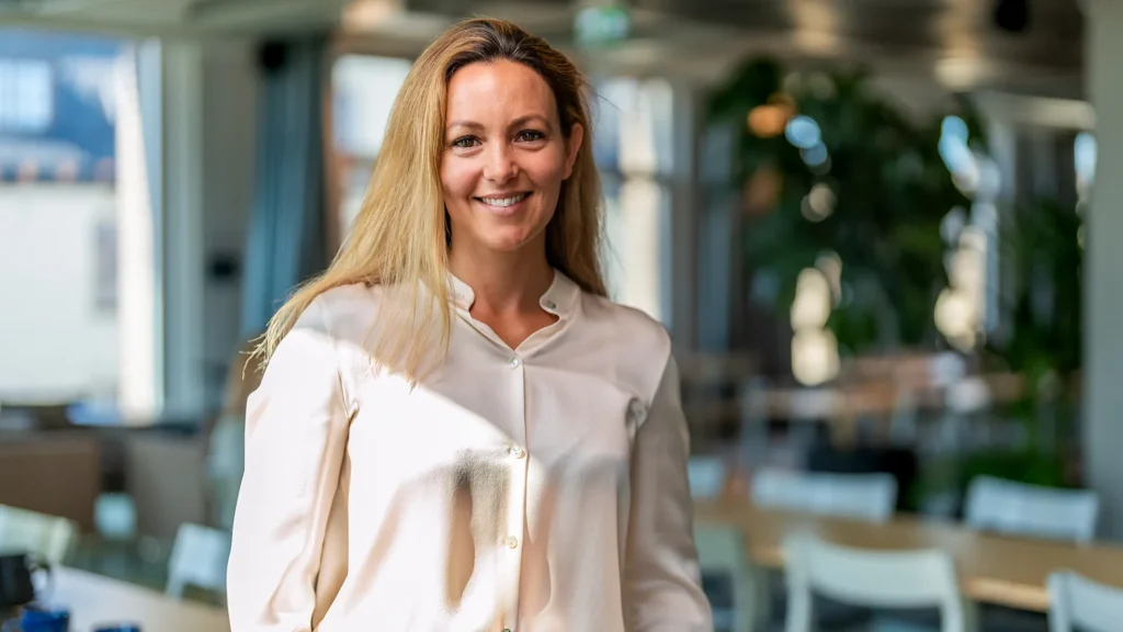En kvinna i kontorsmiljö. Hon tittar in i kameran och ler.