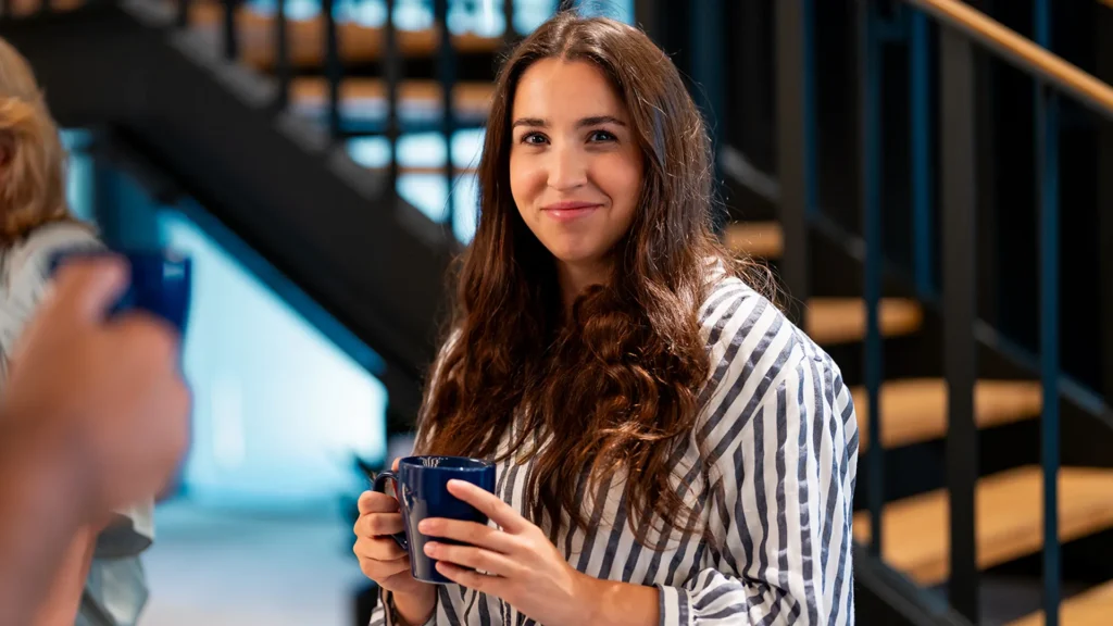 En ung kvinna står vid foten av en trappa. Hon håller en mugg i handen. Hon ler mot kameran.