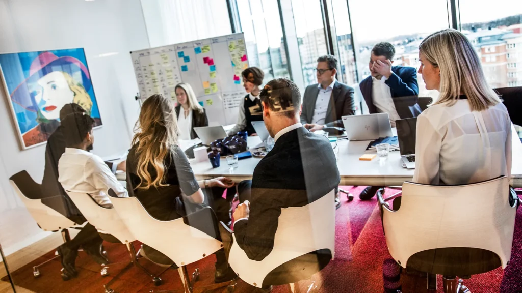 Flera personer sitter runt ett bord i ett kontorsrum. De har en workshop. På en anslagstavla i bakgrunden hänger färgglada postit-lappar.