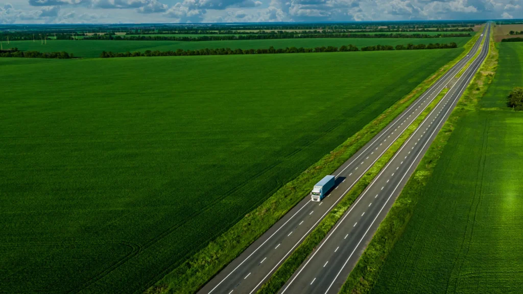 En lastbil kör på en väg som går genom ett grönskande åkerlandskap. Bilden är tagen snett ovanifrån.