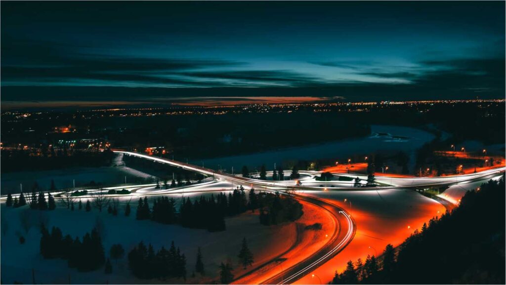 Flygfoto i nattljus av snötäckt trafikplats med slingrande motorvägar och ljusspår från fordon.